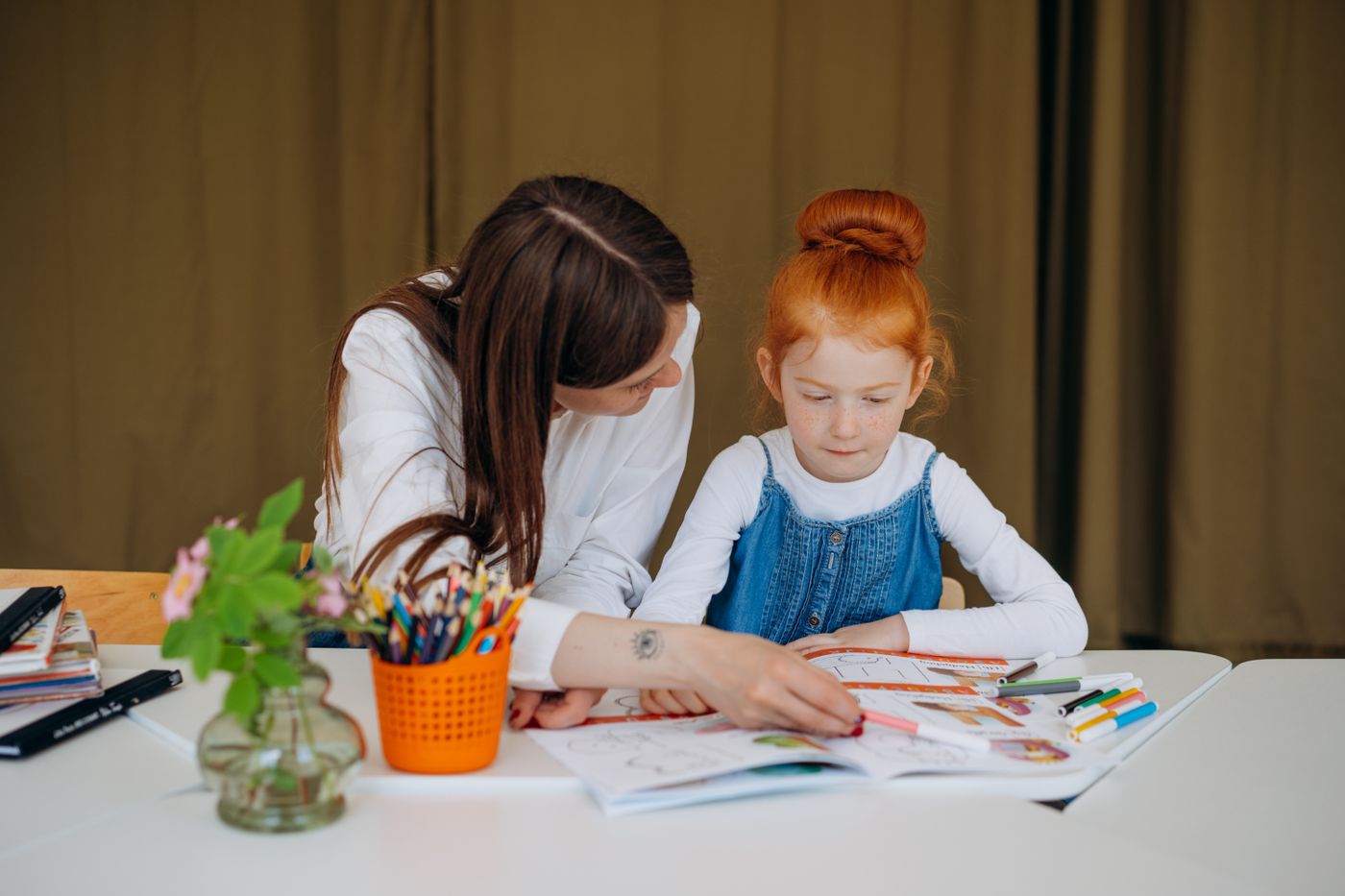 An educator sitting beside a primary-school child, leaning in at her level and working through the page together.